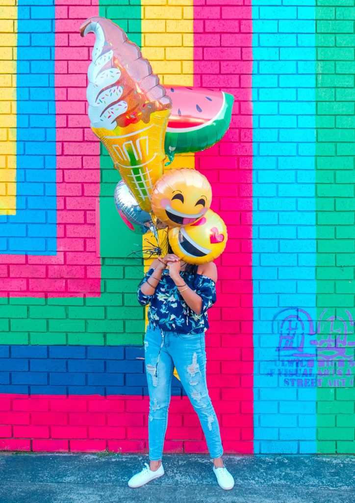 Woman in front of bright mural with emoji balloons in front of her face.